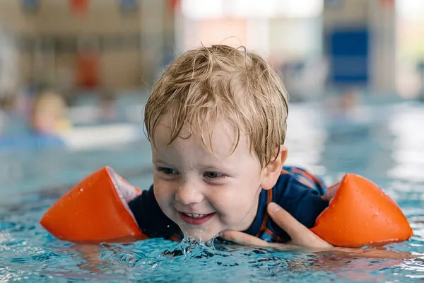 Un bambino con i braccioli avanza a pelo d’acqua, accompagnato dall’istruttrice, durante una lezione genitore-bambino in vasca coperta — Genitore-bambino (2-4 anni)