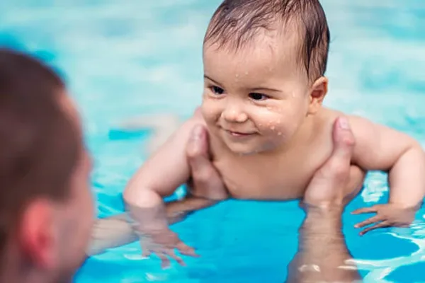 Un bebè sorride a pelo d’acqua appoggiato a un tubolare galleggiante, durante una lezione di acquaticità Acquababy in vasca coperta — Acquababy · Genitore-bebè (0-12 mesi)